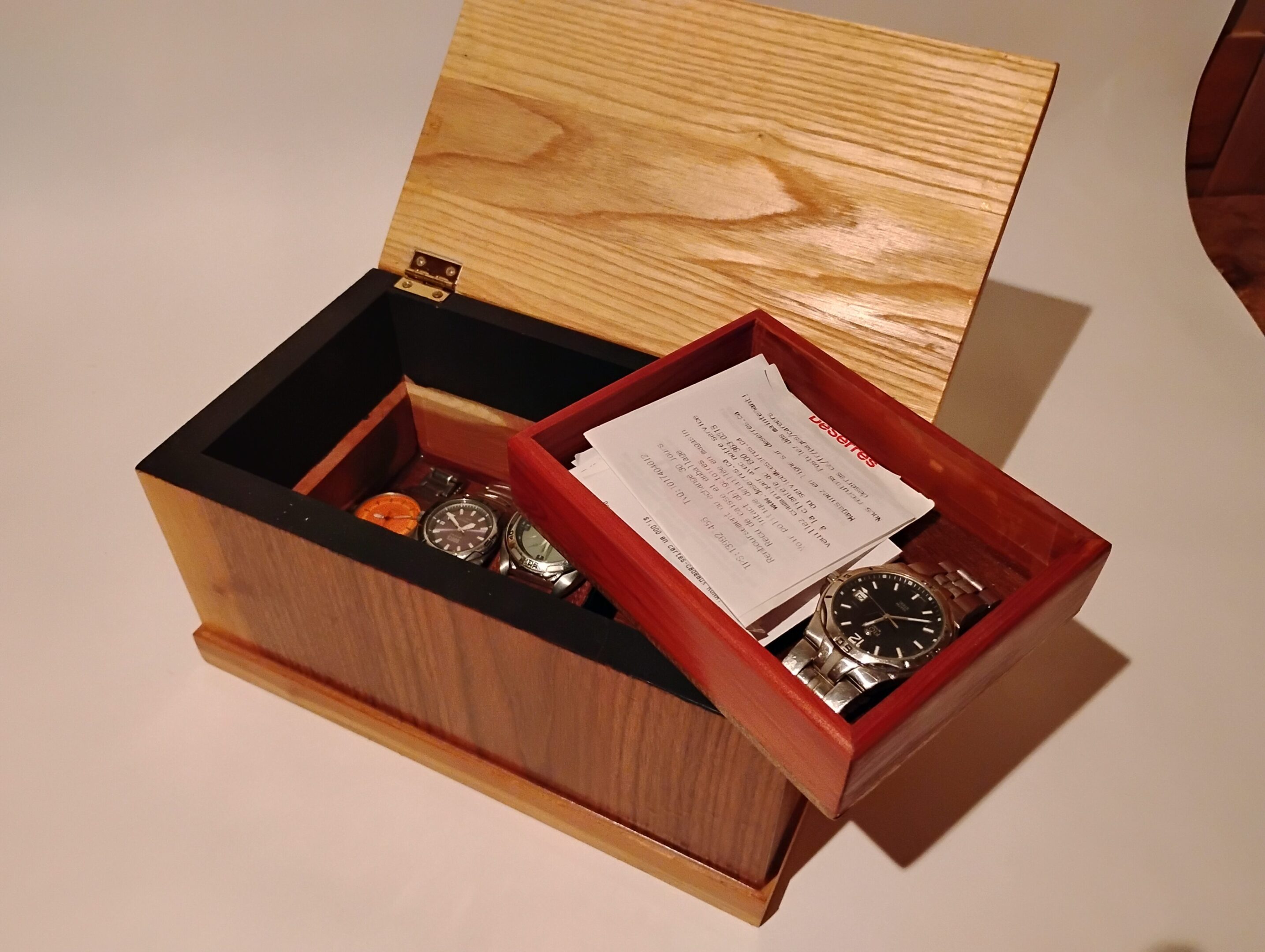 Wooden box with an open lid containing watches and a red tray with papers.